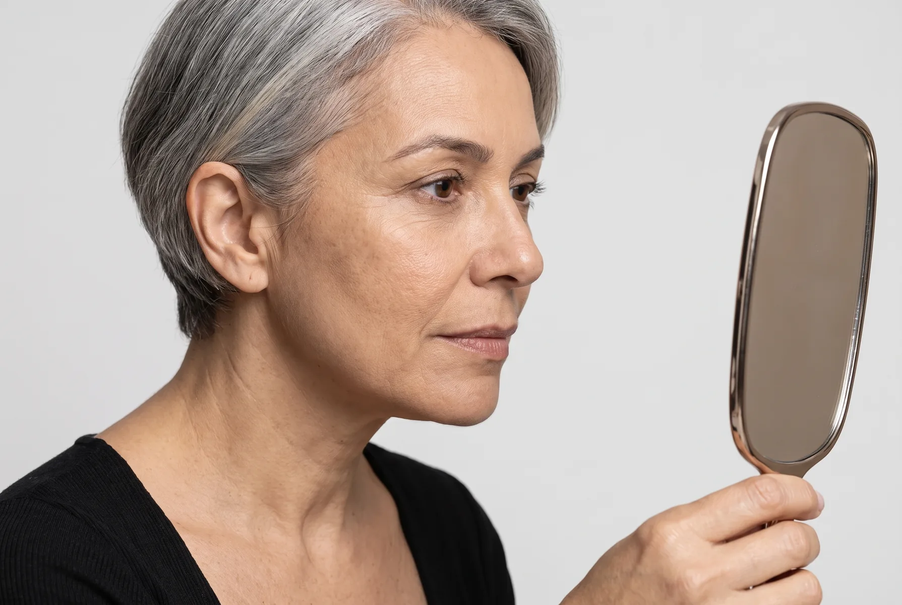Close de uma mulher de 52 anos de pele clara e cabelo grisalho curto, olhando para um pequeno espelho de mão, observando a região dos olhos, pele