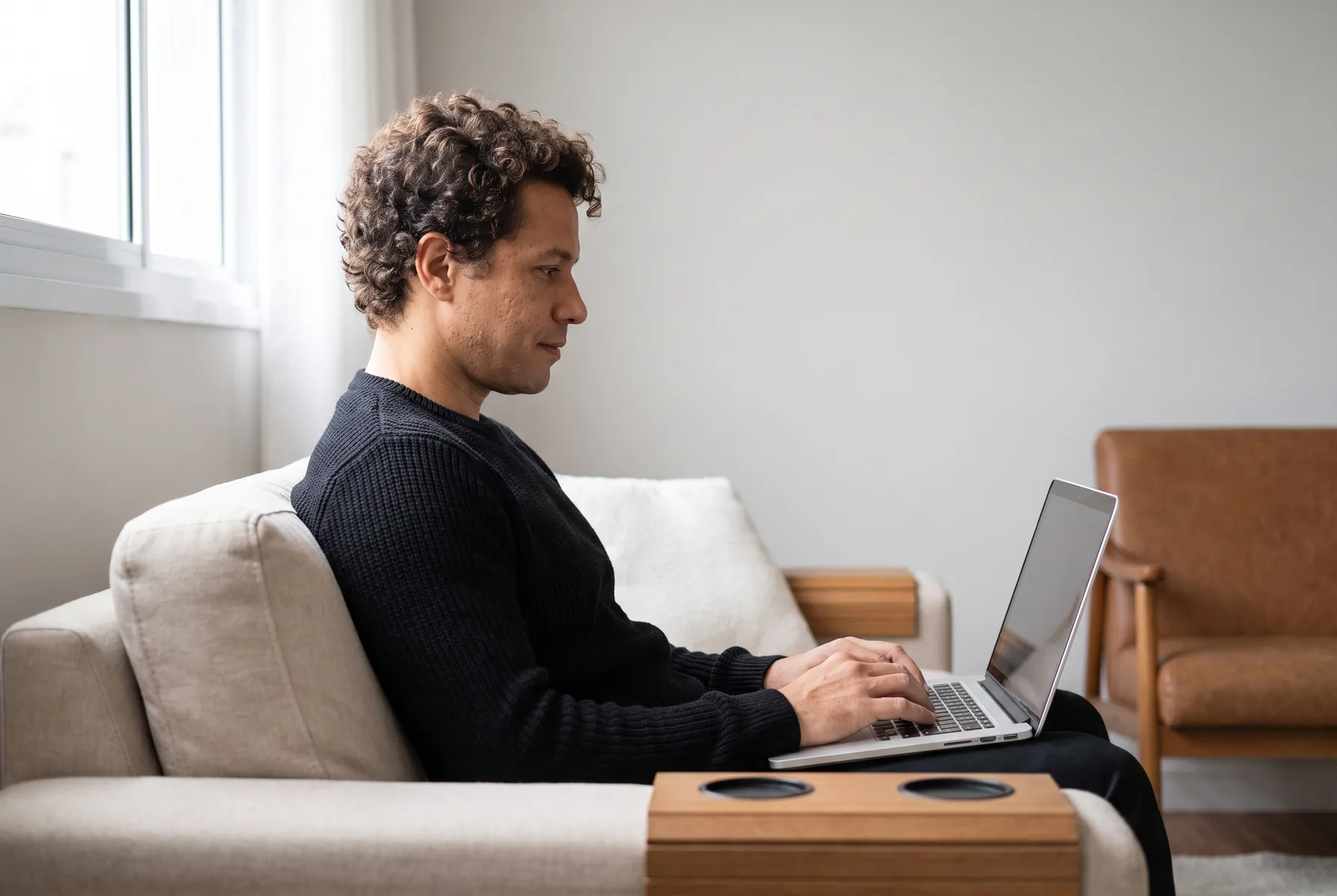 Homem de 34 anos trabalhando em casa no notebook, sentado de forma relaxada no sofá de três quartos