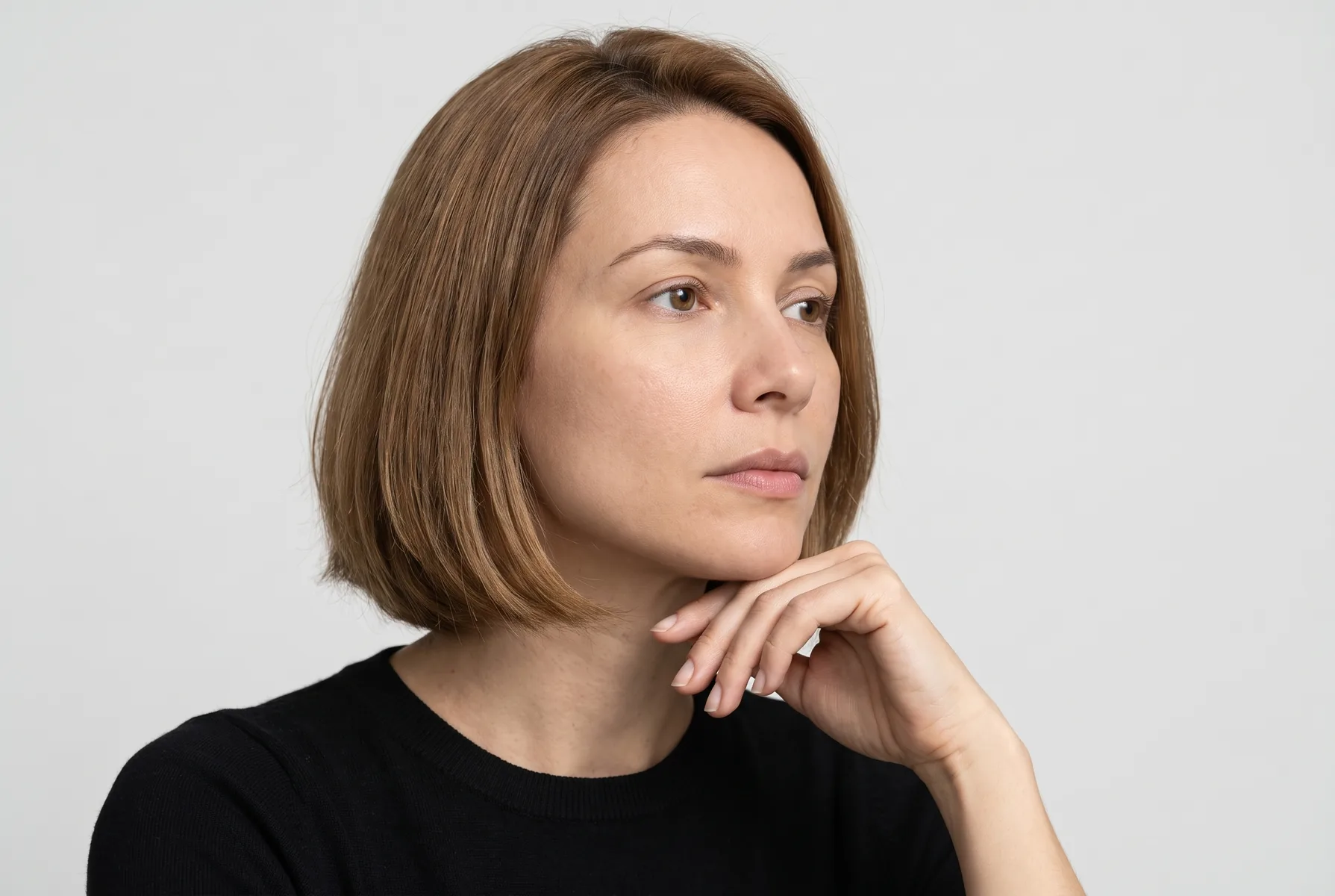Mulher de 36 anos de pele clara, cabelo castanho-claro curto em corte chanel, de três quartos, com a mão apoiada de leve no queixo e o olhar