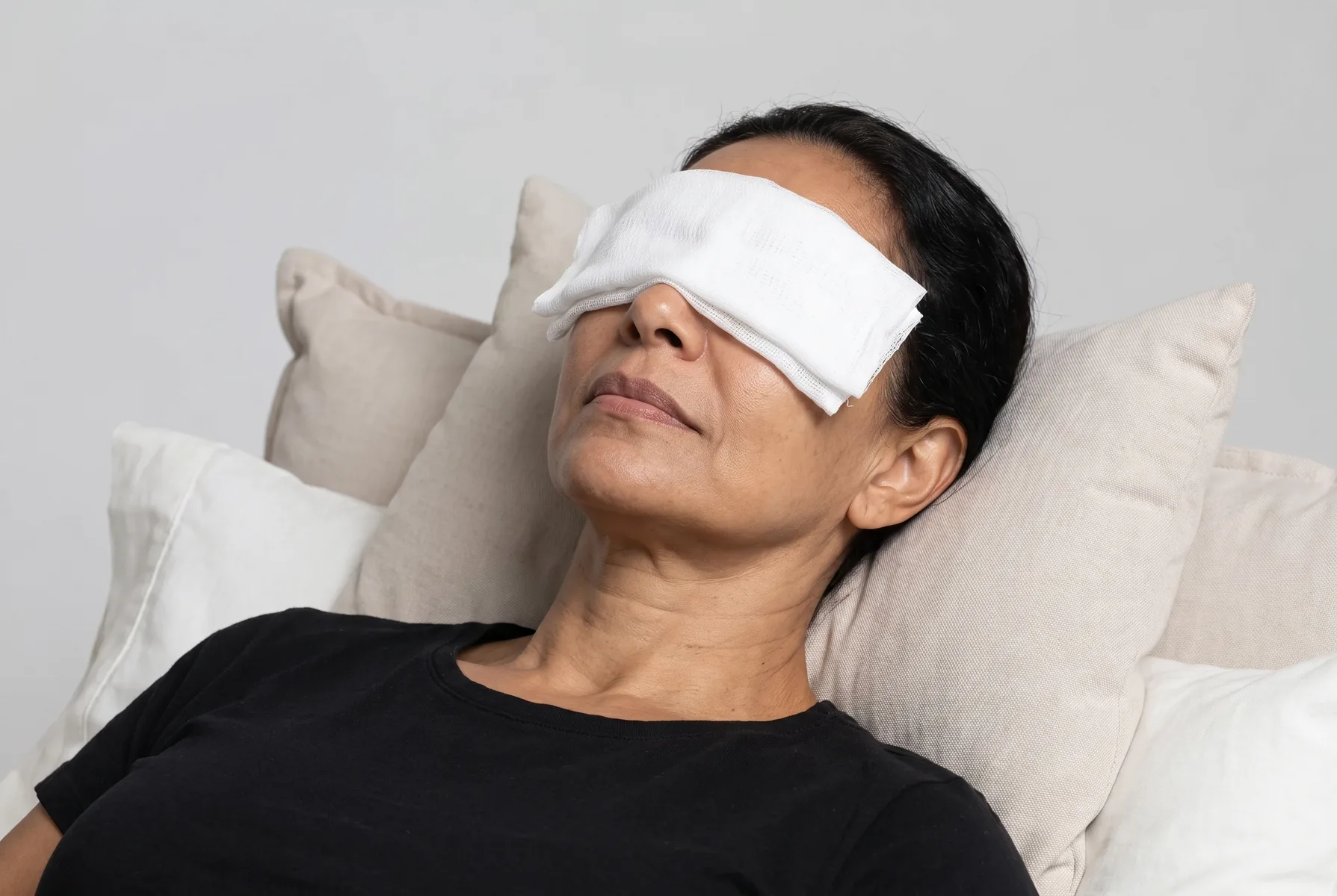 Mulher de 56 anos reclinada em travesseiros claros, com compressas frias de gaze branca sobre os olhos fechados, expressão relaxada