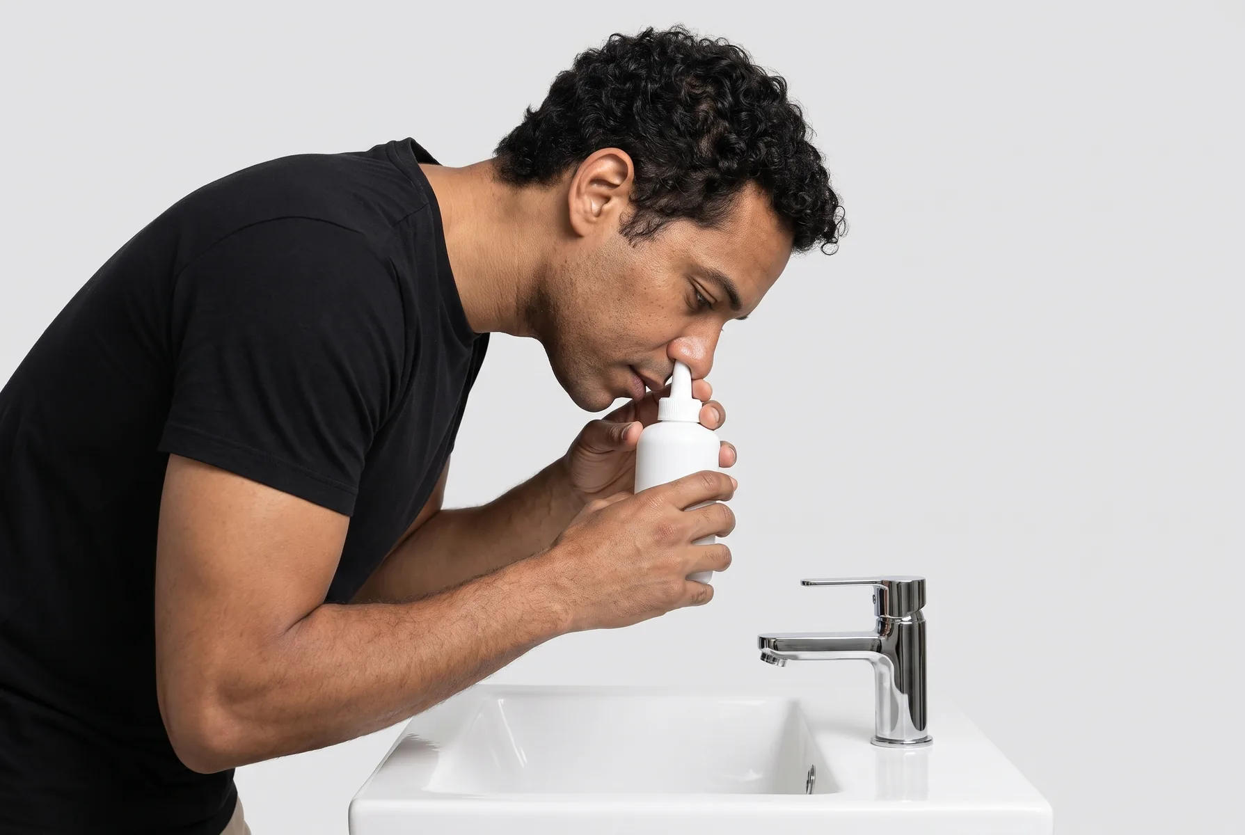 Homem de 36 anos em um banheiro claro e limpo, fazendo lavagem nasal com um frasco simples de soro, inclinado sobre a pia, de