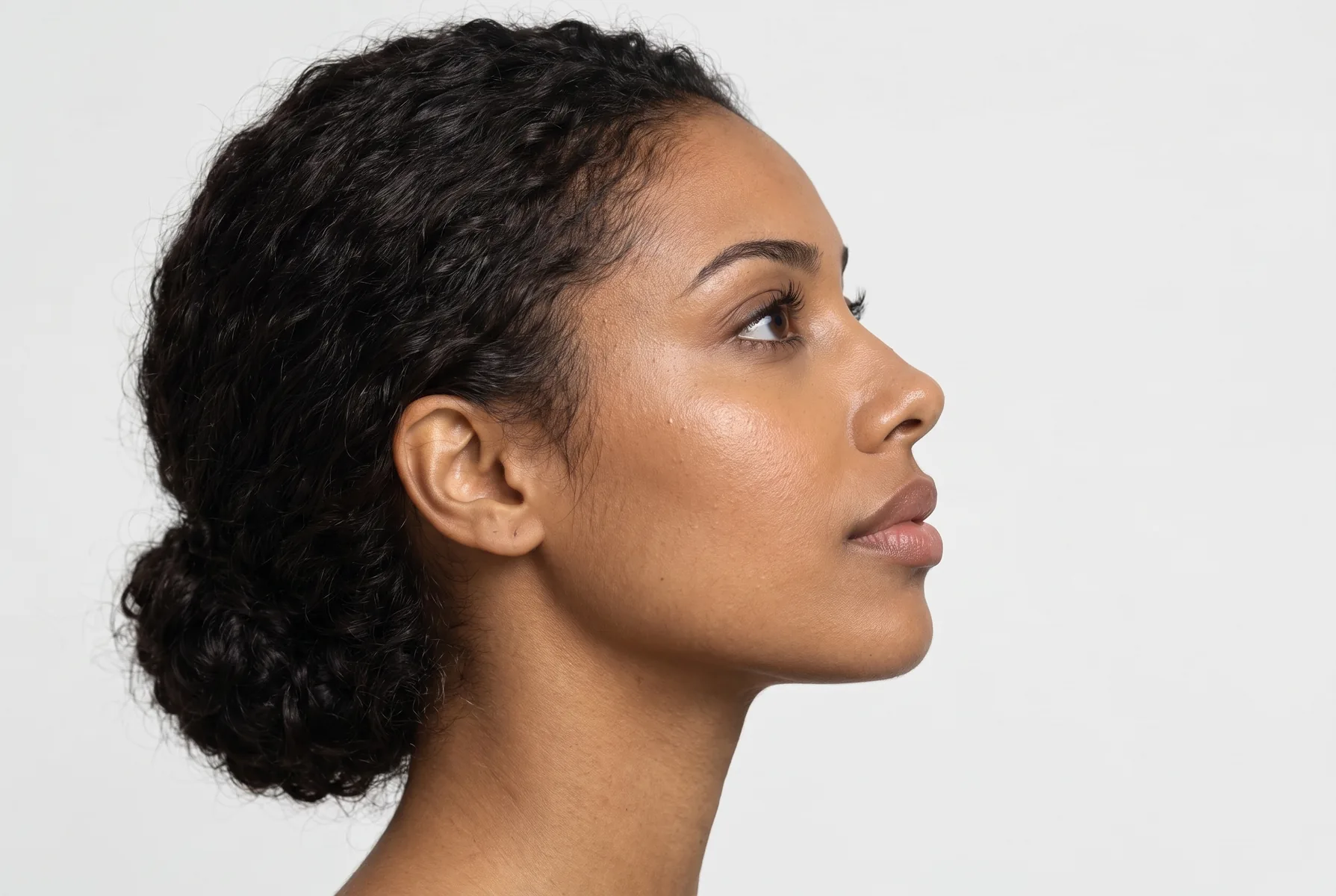 Close de perfil de uma mulher de 28 anos de pele morena clara e cabelo castanho-escuro ondulado preso, enquadrado do olho ao queixo, levemente de