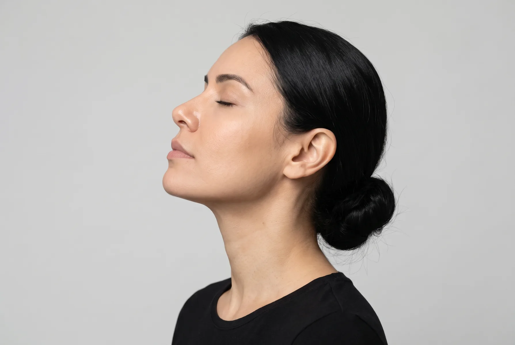 Mulher de 34 anos de pele clara, cabelo preto liso preso em coque baixo, de perfil, olhos fechados, inspirando
